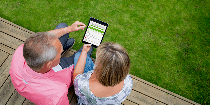A man working on a  laptop and a woman on the phone in a garage with garden tools in the background.
